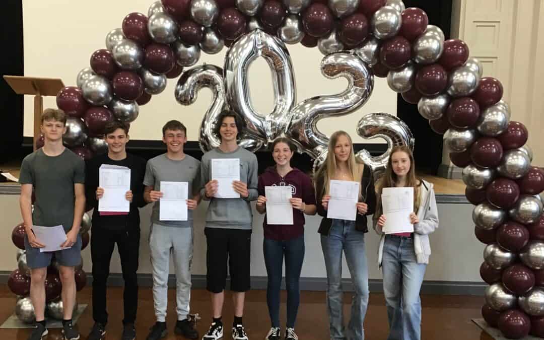 Hazel Grove High School students collect their results on GCSE results day 2023. 7 students hold their results and smile under a balloon arch.