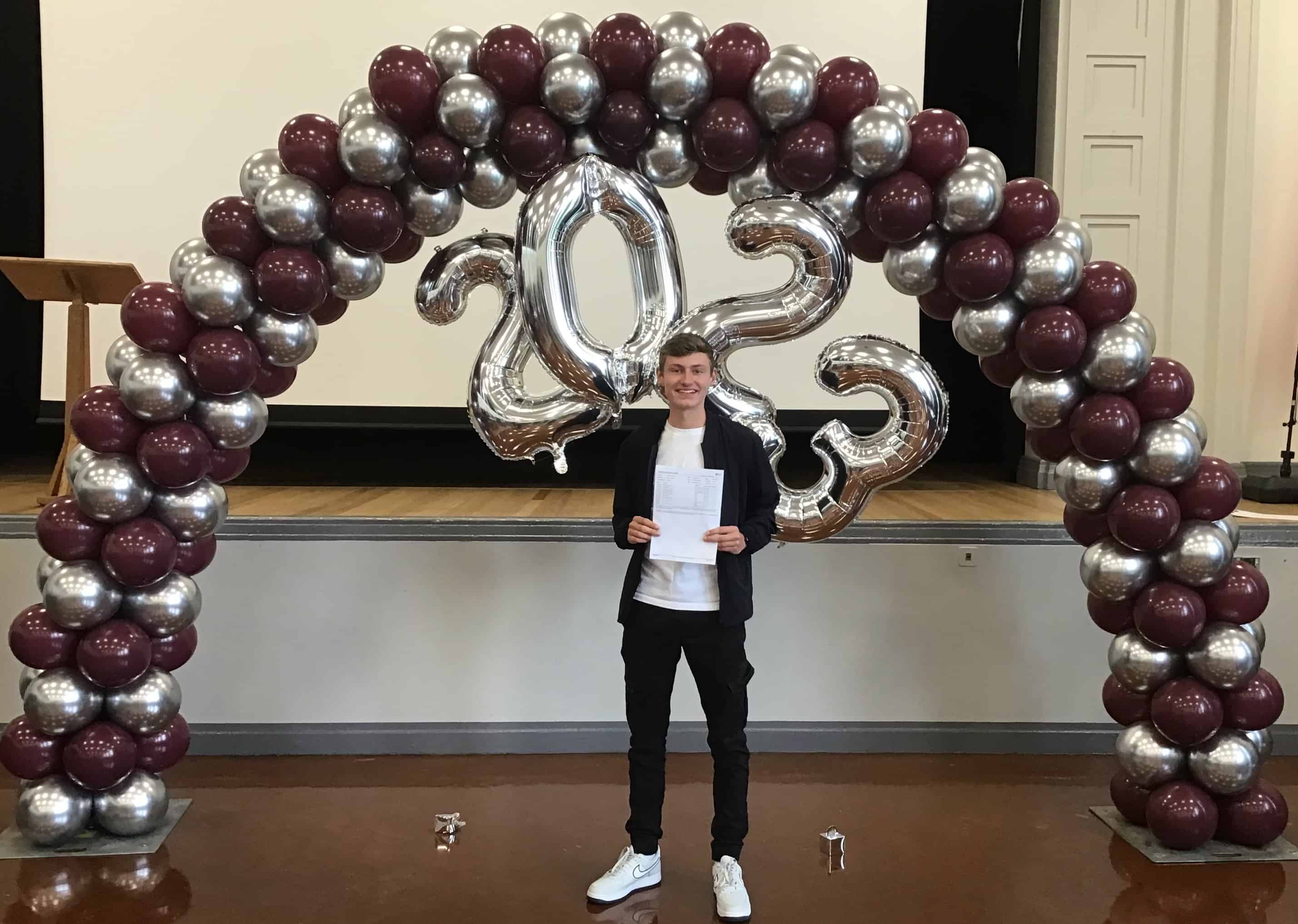 Elliot, Head Boy of Hazel Grove High School, smiles under a balloon arch holding up his results on GCSE results day.