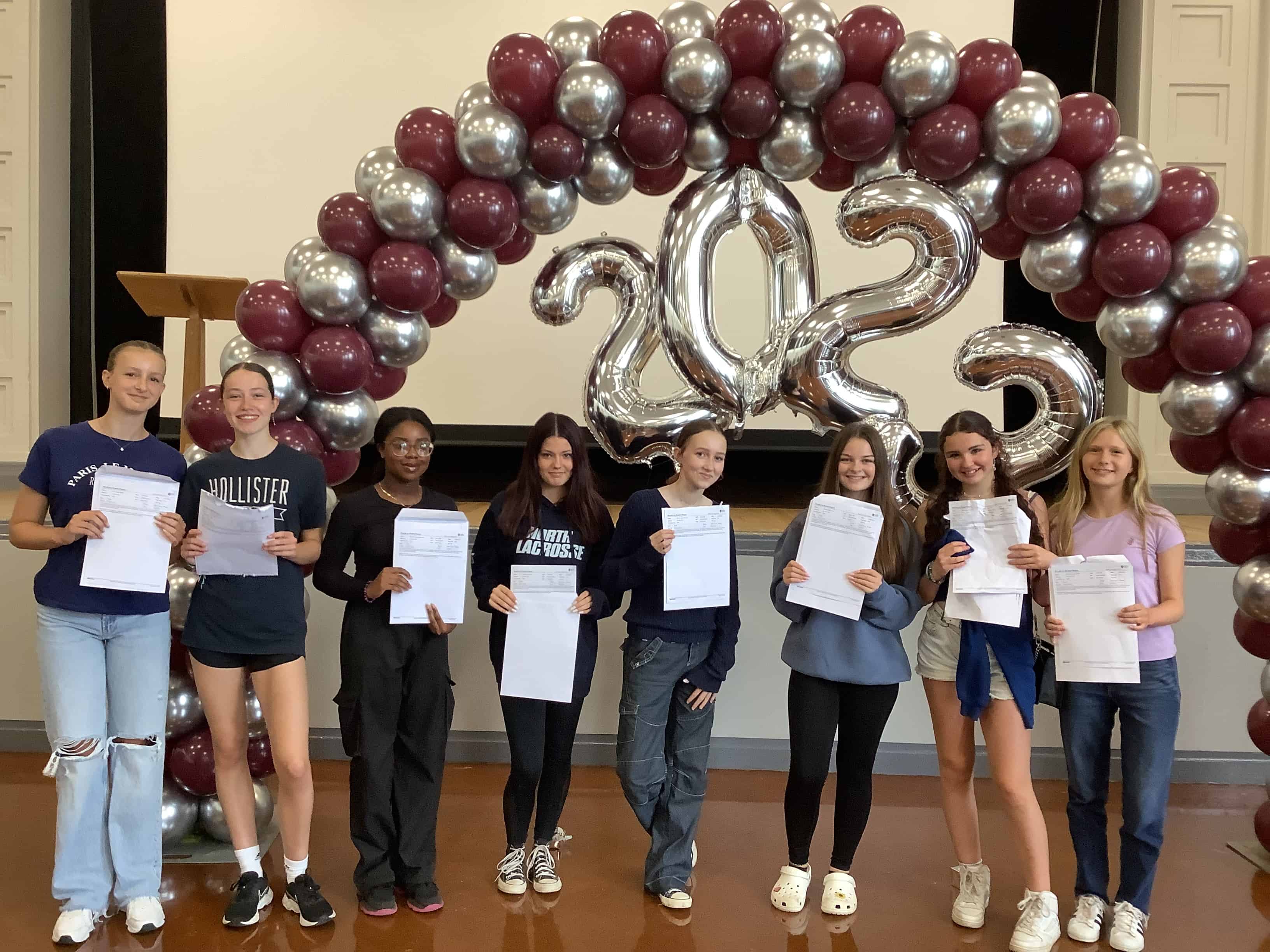 Eight Year 9 students from Hazel Grove High School smile holding their MFL results under a balloon arch at GCSE results day 2023.