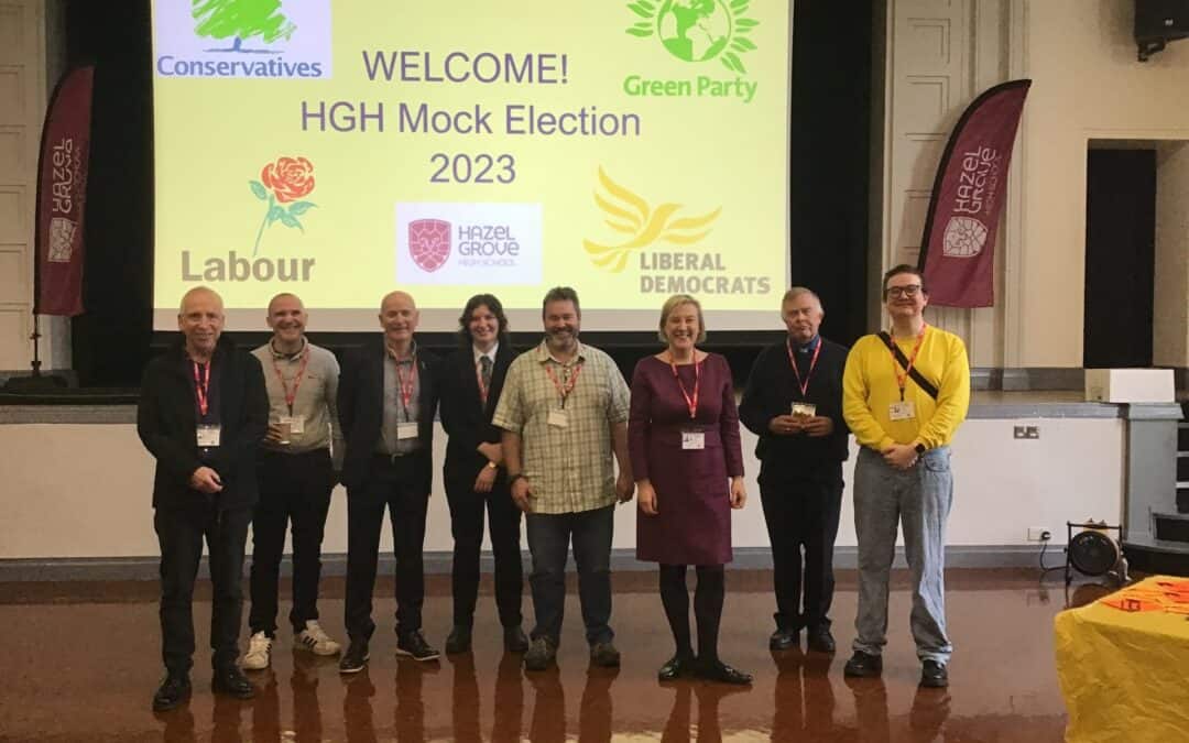 Local representatives from different political parties stand together at Hazel Grove High School in front of a screen which says: WELCOME! HGHS Mock Election 2023