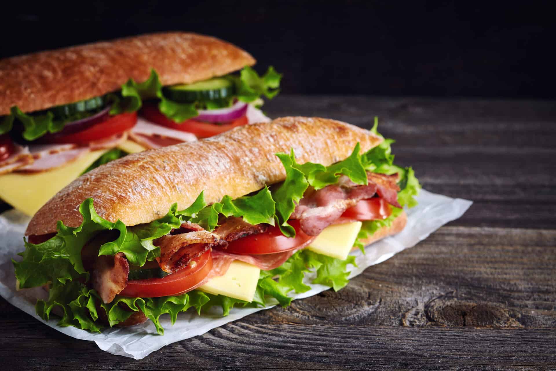 Two fresh submarine sandwiches with ham, cheese, bacon, tomatoes, lettuce, cucumbers and onions on dark wooden background. An example of school lunches at Hazel Grove High School Canteen for illustrative purposes.